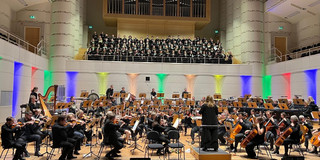 Heinke Kirzinger dirigiert die Zugabe. Vorne sitzt das Orchester. Auf der Empore steht der Chor.